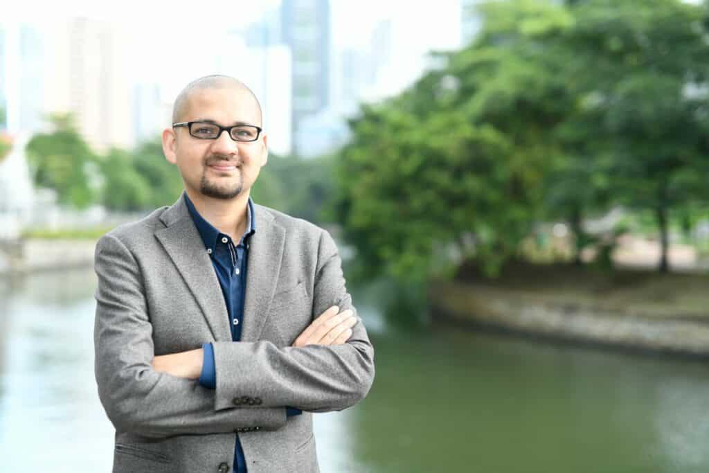 A portrait of Dr. Chris Vas with his arms crossed in front of a river with a city skyline, wearing a grey suit and black framed glasses.