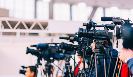 A row of video cameras at a press event, against a commercial background.